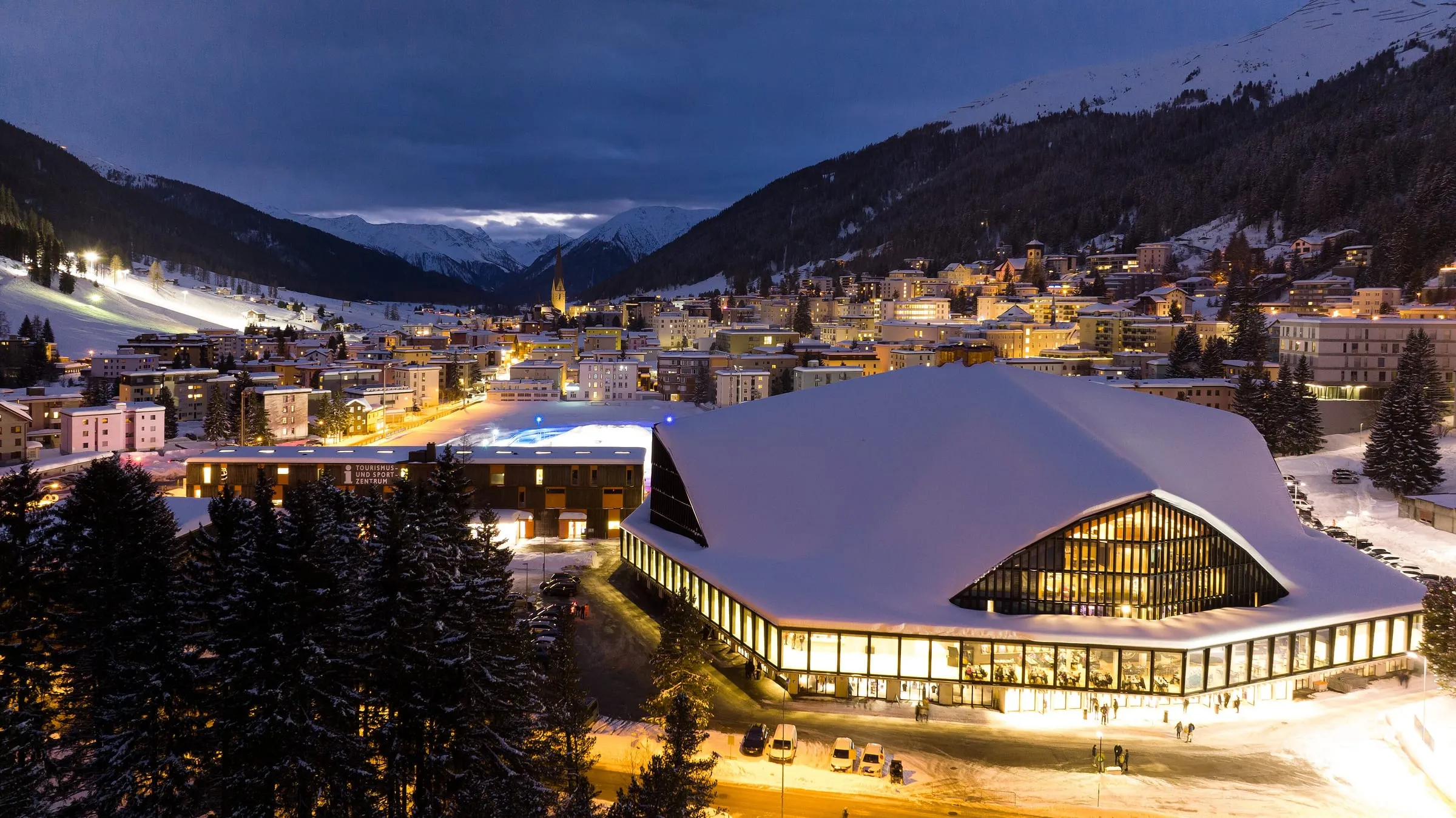 The Davos ice stadium lit up at dusk, with the town and snowy mountains behind