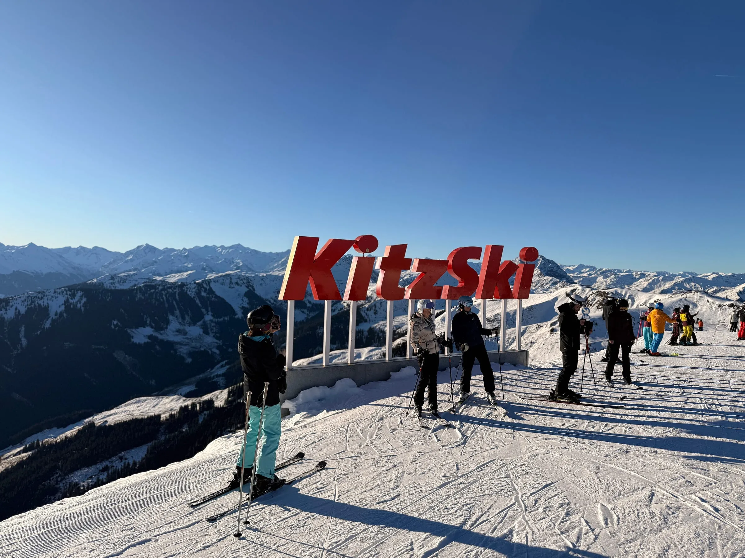 Skiers beside the KitzSki sign at the Jufenalm top station on a sunny winter day