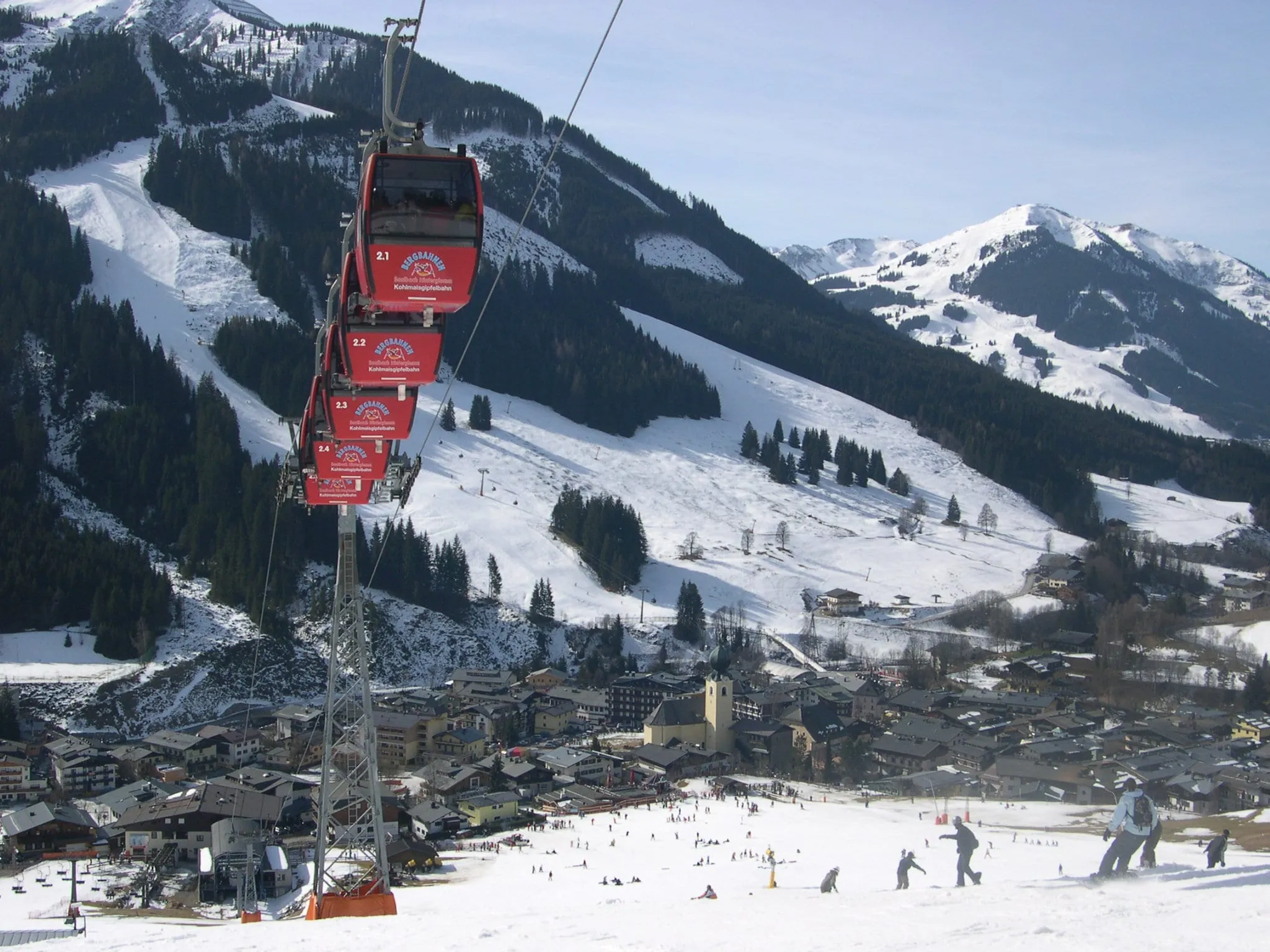 Red Kohlmaisbahn gondolas above Saalbach village and its ski slopes in winter, with the Schattberg and Zwölferkogel behind