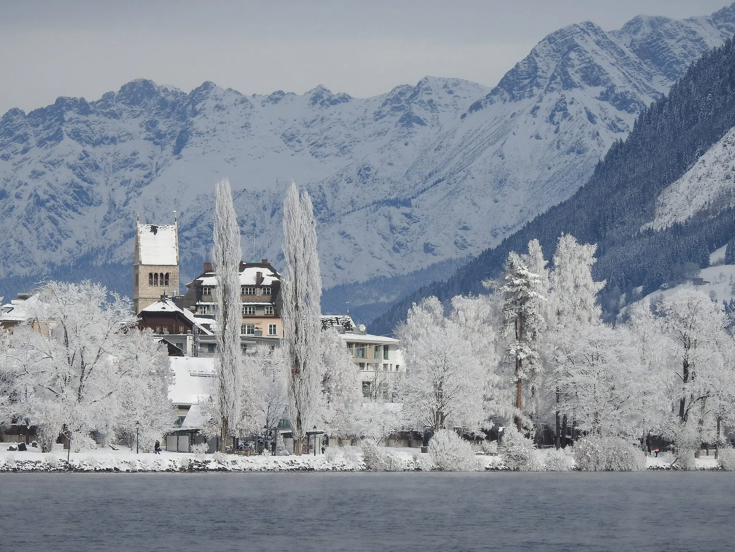 Zell am See's lakefront and church tower in winter, with frost-covered trees and snowy mountains behind
